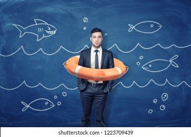 A young businessman stands inside an orange life buoy on a blue background with chalk waves and fish. Business insurance. Blue ocean strategy. Big company in small town.