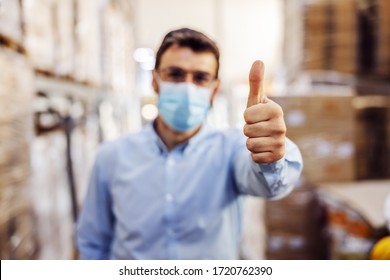 Young Businessman With Sterile Protective Mask On Standing In Warehouse And Showing Thumbs Up. Protection From Corona Virus/ Covid-19. Selective Focus On Hand.