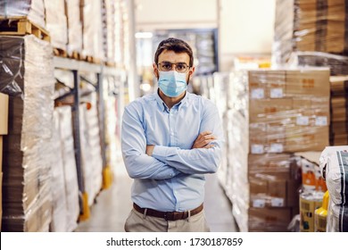 Young businessman with sterile protective mask on standing in warehouse with arms crossed. Protection from corona virus/ covid-19. - Shutterstock ID 1730187859