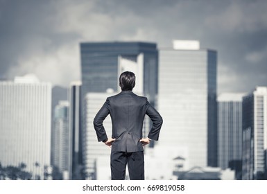 Young Businessman In A Strong Standing Pose Looking Over The City With Dark Storm Clouds Hanging Over. Conquer Your Fears, Success And Determination Concept.  