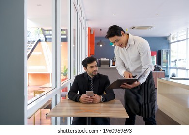 Young Businessman In Suit Listening To The Waiter While Holding Mobile Phone. Asian Waiter In Apron Pointing Finger At Restaurant Menu. Recommend Chef Choice Menu To The Customer.