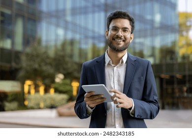 Young businessman in suit using tablet outside modern glass office building. Confident professional smiling working on digital device. Concept of business, technology, success, and entrepreneurship. - Powered by Shutterstock