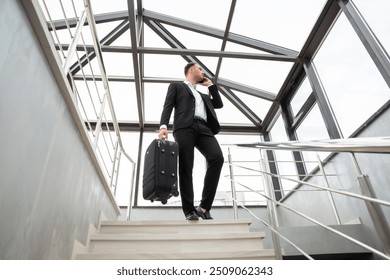 Young businessman with suitcase in stylish suit talking by phone while walking by stairs in modern business building - Powered by Shutterstock