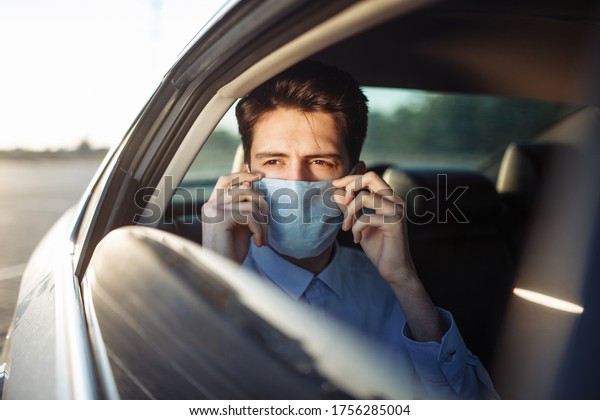Young businessman takes a taxi and looks out of\
the car window adjusting sterile medical mask. Man sits on the back\
seat of taxi and takes a ride during coronavirus pandemic. Social\
distance concept.