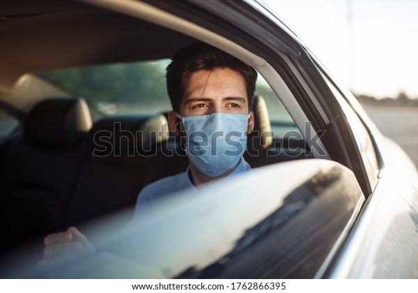 Young businessman takes a taxi and looks out of\
the car window wearing sterile medical mask. A man sits on the back\
seat of taxi and takes a ride during coronavirus pandemic. Social\
distance concept.