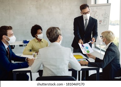 Young Businessman Talking To His Coworkers While Analyzing Charts During A Meeting In Board Room. All Of Them Are Wearing Protective Face Masks Due To COVID-19 Epidemic. 