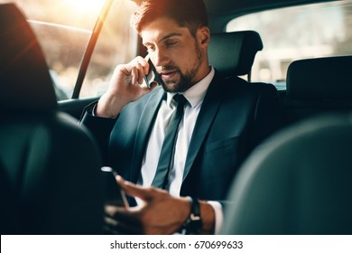 Young Businessman Talking On Mobile Phone And Using Tablet Pc While Sitting On Back Seat Of A Car. Caucasian Male Business Executive Travelling By A Taxi And Looking At Digital Tablet.