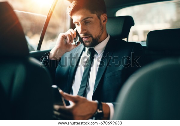 Young\
businessman talking on mobile phone and using tablet pc while\
sitting on back seat of a car. Caucasian male business executive\
travelling by a taxi and looking at digital\
tablet.