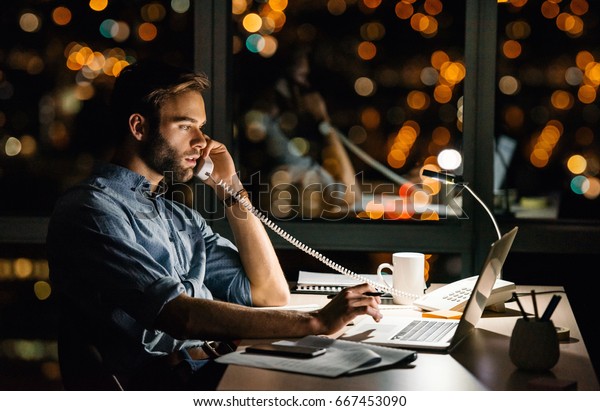 Young businessman talking on the phone and using a\
laptop at his office desk late t night in front of windows\
overlooking the city 