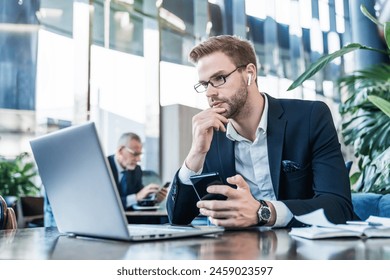 Young businessman trader crypto investor IT-worker in earphones using holding mobile phone and looking on laptop while working at office hall modern coworking. Business concept - Powered by Shutterstock