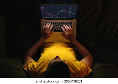 Young businessman trader investor using stockmarket app on laptop for cryptocurrency financial market analysis on sofa at home, trading data index chart graph on laptop screen. Top view