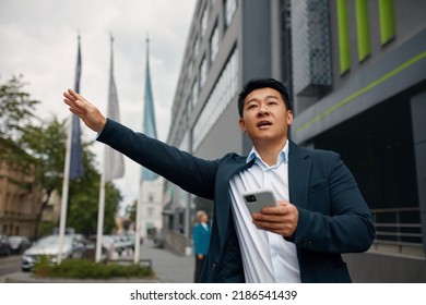 Young Businessman Waiting For Taxi Cab On The Street In The City. Asian Man Stop The Car On City Street. Guy Standing On The Road Waving His Hand Outside 