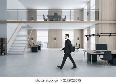Young businessman walking in modern two floors wooden and concrete duplex office interior with panoramic city view, furniture and daylight. Worker and ceo concept