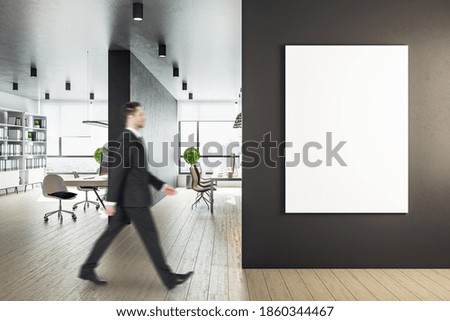 Young businessman walking in workplace interior with computers and blank banner on wall. Workplace and worker concept. Mock up.
