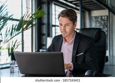 Young businessman wearing black suit, sitting to table with laptop. Office manager working in modern office with glass windows blurred background, glass of water on the table, concept of work - Powered by Shutterstock