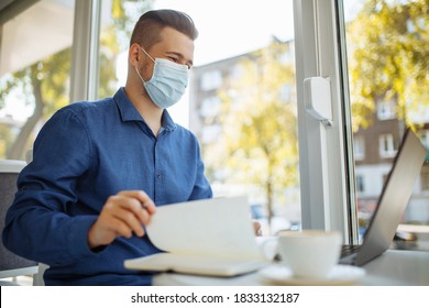 Young businessman wearing a medical mask sits in a cafe with a laptop working and taking notes. Coronavirus pandemic, distant work and business concept. - Shutterstock ID 1833132187