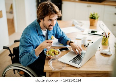 Young Businessman In Wheelchair Eating While Surfing The Net On Laptop On Lunch Break At Home. 