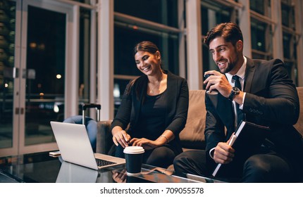 Young Businessman And Woman Waiting At Airport Lounge And Looking At Laptop. Business Travelers Waiting For Their Flight At Airport Terminal.