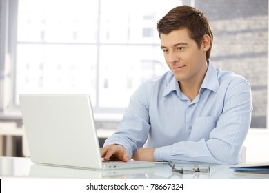 Young businessman working in office, sitting at desk, looking at laptop computer screen, smiling.?