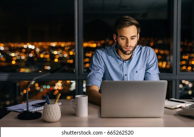 Young Businessman Working On A Laptop At His Office Desk Late Into The Night In Front Of Windows Overlooking The City 