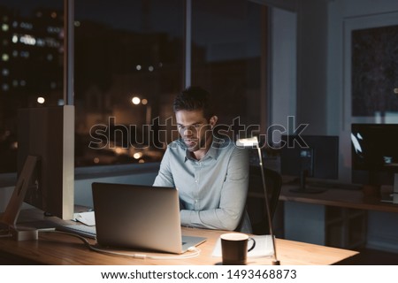 Young businessman working overtime alone at his desk in an office late at night with city lights glowing in the background