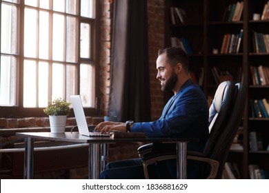 A young businessman works on a laptop in a luxurious office in the library. A bearded, cheerful man in a blue suit at a Desk is typing on a keyboard and smiling - Shutterstock ID 1836881629