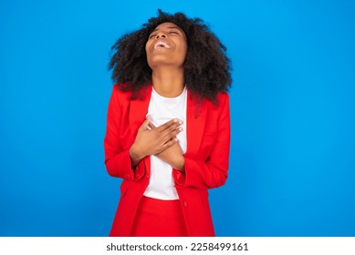 young businesswoman with afro hairstyle wearing red over blue background expresses happiness, laughs pleasantly, keeps hands on heart - Shutterstock ID 2258499161