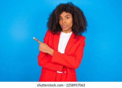 young businesswoman with afro hairstyle wearing red over blue wall smiling broadly at camera, pointing fingers away, showing something interesting and exciting. - Powered by Shutterstock