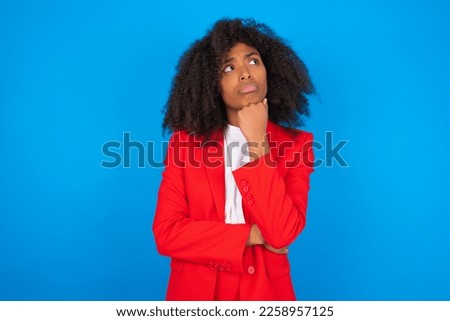 young businesswoman with afro hairstyle wearing red over blue wall with hand under chin and looking sideways with doubtful and skeptical expression, suspect and doubt.