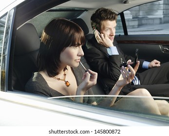 Young Businesswoman Applying Lip Gloss And Businessman Using Cell Phone In Car