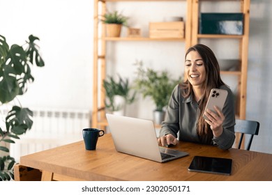 Young businesswoman with beautiful smile in home office holding mobile phone working on laptop sitting at desk.