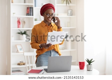 Young businesswoman cheerful african american lady with red turban on her head working at office, standing by workdesk with laptop, holding documents, having phone conversation, copy space