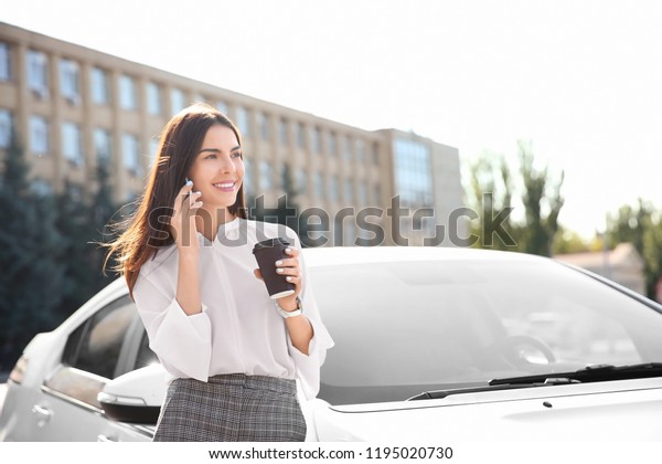 Young businesswoman with coffee talking on mobile\
phone near car