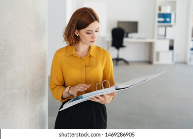 Young businesswoman consulting a large handheld file as she pauses alongside a wall in the office