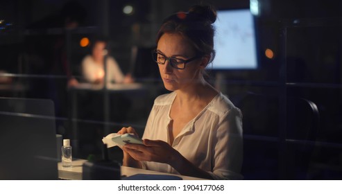 Young Businesswoman Disinfecting Smartphone With Sanitizer Gel Working Late In Office. View Through Window Of Female Executive Cleaning Mobile Phone With Antiseptic Sitting At Workplace At Night