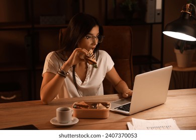 Young businesswoman eating tasty sandwich in office at night - Powered by Shutterstock