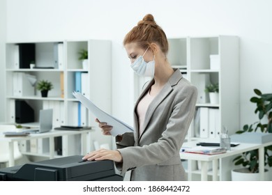 Young Businesswoman In Elegant Grey Suit And Protective Mask Looking Through Paper While Standing By Xerox Machine In Office Environment