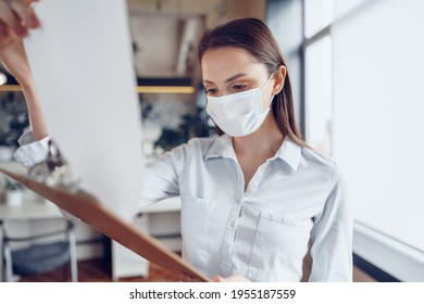 Young Businesswoman In Face Mask Standing In Office And Holding Clipboard With Documents