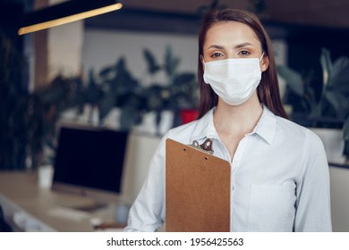 Young Businesswoman In Face Mask Standing In Office And Holding Clipboard With Documents