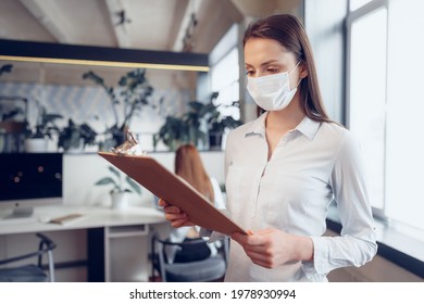 Young Businesswoman In Face Mask Standing In Office And Holding Clipboard With Documents