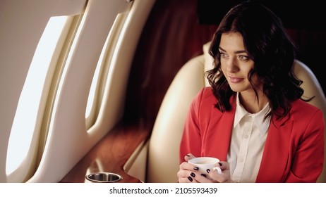 Young businesswoman holding coffee and looking at windows in private jet