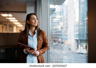 Young businesswoman holding digital tablet and day dreaming while looking through the window from her office. Copy space.