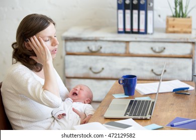 Young businesswoman holding newborn crying babe while sitting with laptop and touching head with pained expression. Career mom suffering from migraine after working on pc and nursing crying child