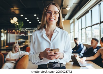 Young businesswoman holding a smartphone in a co-working space. Happy young businesswoman smiling at the camera while standing in a modern workplace. Female entrepreneur sending a text message.