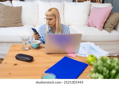 Young businesswoman holding smartphone while working with laptop at home office desk, using cross platform responsive design apps for project analysis, analyzing cloud data statistics. Freedom concept - Powered by Shutterstock