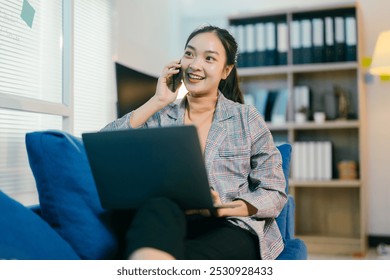 Young businesswoman multitasks, talking on the phone and typing on her laptop in a modern office setting, exuding professionalism and happiness - Powered by Shutterstock