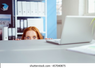 Young businesswoman peering over her desk in the office in wide eyed horror or amazement with just her eyes visible