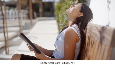 Young businesswoman relaxing on an outdoor wooden bench sitting with her head tilted back and eyes closed while holding a tablet computer  side view