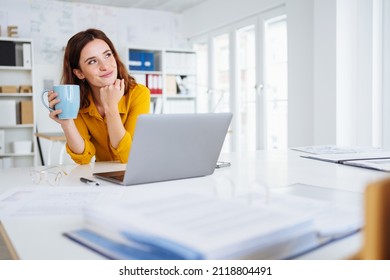 Young businesswoman reliving happy memories during a coffee break sitting at her desk in the office looking up to the side with a smile of contentment and pleasure - Shutterstock ID 2118804491