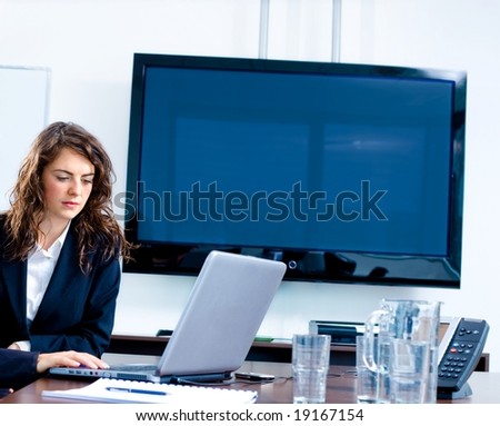 Young businesswoman sitting by meeting table at office in front of a huge blank plasma TV screen and using laptop and phone.